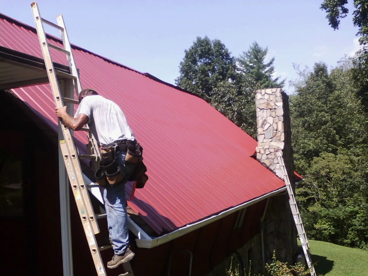 Roofer on a ladder beside a red metal roof and stone chimney — Soffit & Fascia Repair by Roofing Brothers in Weldon